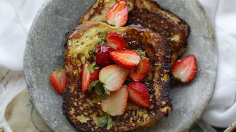 Two slices of vegan French toast on a gray plate, topped with fresh sliced strawberries and drizzled with syrup.